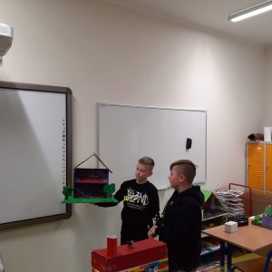 Uczeń klasy czwartej stoi obok tablicy trzymając w dloniach wykonaną przez siebie makietę Akademii Pana Kleksa. Makieta przedstawia przekrój dwupietrowego budynku ze spadzistym dachem. Obok stoi inny uczeń przyglądający się makiecie.