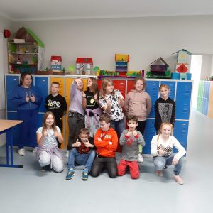 Uczniowie klasy czwartej pozują do zdjęcia na tle kolorowych szkolnych szafek na których ustawiono makiety akademii Pana Kleksa przez nich wykonane.Część uczniów podnosi w górę kciuki.