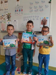 Trzech chłopców z oddzialu przedszkolnego stoi obok siebie na tle klasowej ściany. Chłopcy trzymają w dłoniach przed sobą otrzymane upominki z okazji dnia chłopala - kolorowanki.