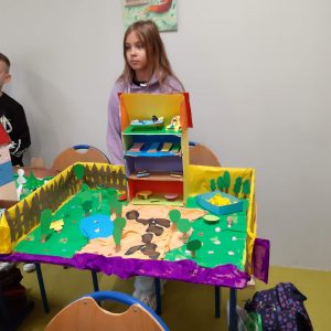 Uczennica klasy czwartej stoi za wykonaną przez siebie makietą Akademii Pana Kleksa. Makieta przedstawia przekrój 3 piętrowego budynku oraz teren w okół niego z drzewami i łąkami.