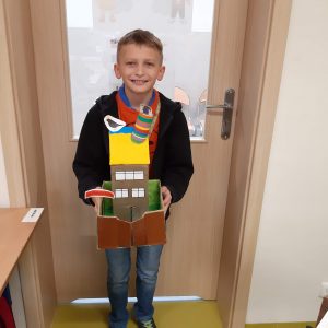 Uczeń klasy czwartej stoi przed drzwiami trzymając w rękach wykonaną przez siebie makietę Akademii Pana Kleksa. Makieta przedstawia wysoki budynek z żółtym dachem i kolorowym kominem, otoczony wysokim płotem.