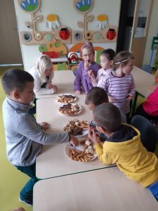 Uczniowie oddziału przedszkolnego stoją przy ustawionych w rzędzie stołach. Na stolikach ustawiono cztery talerze ze słodkościami dla dzieci. Uczniowie jedzą słodycze i rozmawiają ze sobą.