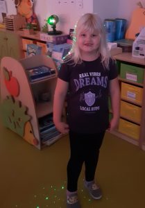 Uczennica oddziału przedszkolnego pozuje do zdjęcia w swojej sali. Na włosach dziewczynki i przy jej stopach świecą się zielone laserowe światełka.