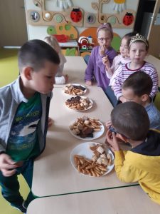 Uczniowie oddziału przedszkolnego stoją przy ustawionych w rzędzie stołach. Na stolikach ustawiono cztery talerze ze słodkościami dla dzieci. Uczniowie jedzą słodycze i rozmawiają ze sobą.