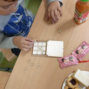 Uczeń siedzi przy stoliku a przed nim leży drewniana gra w kółko i krzyżyk. Chłopiec układa kółka i krzyżyki na drewnianej podkładce.