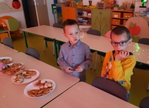 Dwóch uczniów z oddziału przedszkolnego stoi obok siebie przy stole ze słodyczami. Słodycze ułożono na czterach talerzykach a chłopcy je jedzą.