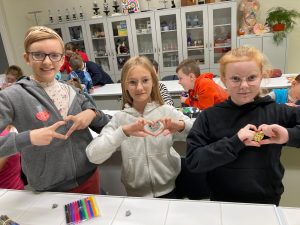 Trzy dziewczynki stoja w rzędzie. Z dłoni wyciagnietych przed siebie formują serca. na ubraniach maja naklejone serdeszka, które własnorecznie pomalowały. W tle widac innych uczniów klasy.