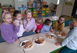 Uczniowie oddziału przedszkolnego stoją przy ustawionych w rzędzie stołach. Na stolikach ustawiono cztery talerze ze słodkościami dla dzieci. Uczniowie jedzą słodycze i rozmawiają ze sobą.