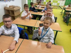 Uczniowie klasy drugiej siedzą parami w ławkach. Chłopcy trzymają w dłoniach otrzymane z okazji dnia chłopaka prezenty - drewniane linijki.