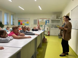 Uczniowie siedza przy stolikach. Na przeciwko stoi Pani bibliotekarka trzymająca książkę.