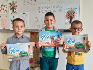 Trzech chłopców z oddzialu przedszkolnego stoi obok siebie na tle klasowej ściany. Chłopcy trzymają w dłoniach przed sobą otrzymane upominki z okazji dnia chłopaka - kolorowanki.