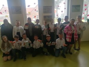 Uczniowie klasy trzeciej pozują do wspólnego zdjęcia. Chłopcy trzymają w dłoniach prezenty otrzymane z okazji dnia chłopaka - kubki i łudeczki z papieru.