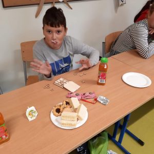 Uczeń siedzi przy stoliku. W buzi ma lizaka, dłoniami macha w stronę aparatu. Na stoliku przed nim stoi talerzy ze słodkościami oraz rozłożona gra w kółko i krzyzyk - prezent z okazji dnia chłopaka.