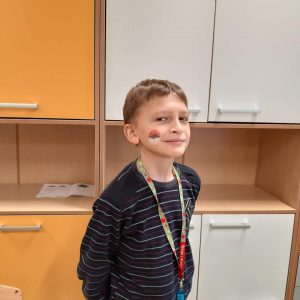 Uczeń pozuje wyprostowany do zdjęcia. Na policzku namalowaną ma biało- czerwoną piłeczkę