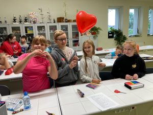 Cztery uczennice stoją za szkolnym stolikiem. Dziewczynki mają przyklejone na ubraniach pokolorowane przez siebie serduszka. Jedna z dziewczynek trzyma balonik w kształcie serca a inna układa przed sobą dłonie formujac serce.