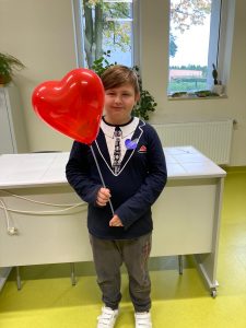 Uczeń pozuje do zdjęcia trzymając w dłoniach czerwony balonik w kształcie serce. Chłopiec ma na ubraniu naklejone serduszko które sam pokolorował.