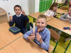 Dwóch uczniów klasy drugiej siedzi przy wspólnej ławce. W dłoniach trzymają prezenty z okazji dnia chłopaka - drewniane linijki i lizaki. W tle widać siedzące w kolejnej ławce dwie uczennice.