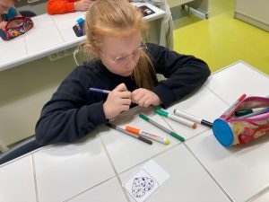 Uczennica siedzi przy szkolnym stoliku. W reku trzyma kolorowy pisak. Na stoliku przed nia leżą inne pisaki oraz karteczka z nadrukowanym sercem.