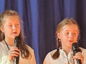Dwie dziewczynki śpiewają do mikrofonów, które trzymaja w dłoniach. Na zdjęciu widać twarze dziewczynek.