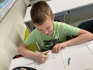 Uczeń siedzi przy szkolnej ławce. W dłoniach ma kolorowy pisek a przed nim lezy karteczka z nadrukowanym sercem.