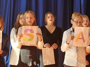 Zdjęcie uczniów klasy szóstej podczas występu artystycznego. Na pierwszym planie widać dwójkę uczniów trzymających karteczki z literami S i A w pomiędzy nimi w kolejnym rzędzie widać uczennicę w czarnej sukience która ma otwarte usta.
