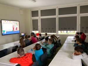 Uczniowiesiedzą przy szkolnych stolikach i ogladają wyświetlona na tablicy multimedialnej grafikę.