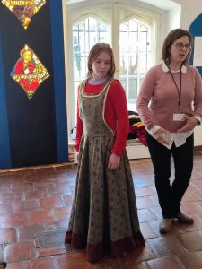Uczennica klasy piątej stoi w stroju mieszczańskim z lekko spuszczoną głową. Obok niej stoi Pani będąca przewodnikiem po muzeum.