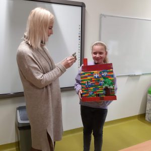 Uczennica klasy czwartej stoi obok nauczycielki. W rękach trzyma wykonana przez siebie makietę akademii Pana Kleksa - kolorowy budynek z czerwonym kominem.