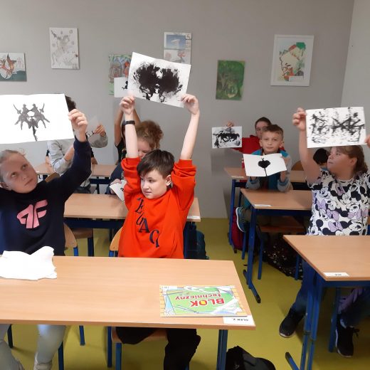 Uczniowie klasy czwartej siedzą w ławkach. W górę podnoszą kartki z wykonanymi przez siebie kleksami.