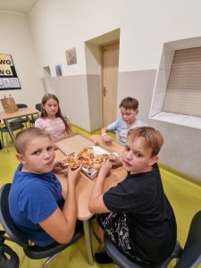 Na szkolnej stołówce czworo uczniów klasy piątej siedzi przy wspólnym stoliku. Na stole lezy pudełko z pizzą. Dwóch chłopców je pizze.