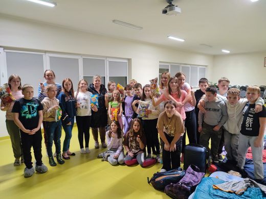 Uczniowie klasy piatej pozują do zdjęcia grupowego podczas nocowania w szkole. Obok uczniów leżą przygotowane do spania materace.