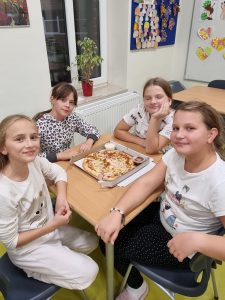 Na szkolnej stołówce cztery dziewczynki siedzą przy wspólnym stoliku, na którym leży pizza. Dziewczynki uśmiechają się do zdjęcia