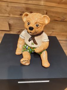 Zdjęcie eksponatu Muzeum Etnograficznego w Toruniu. Na wystawie miś ubrany w białą koszulę i zielone spodenki w kolorowe kropki.