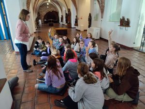 Uczniowie klasy piątej siedzą na podłodze toruńskiego muzeum. część z nich podnosi w górę rękę. Na przeciwko uczniów stoi Pani przewodnik .