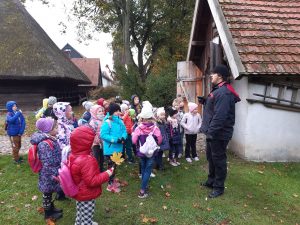 Uczniowie oddziału przedszkolnego stoją przed murowanym domkiem ustawionym na terenie Muzeum Etnograficznego. Obok domku stoi Pan przewodnik. Dzieci odwrócone są bokiem do zdjęcia.