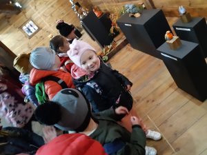 Uczniowie oddziału przedszkolnego podczas zwiedzania wystawy w Muzeum Etnograficznym. Dzieci przyglądają się eksponatom. Dziewczynka stojąca z przodu uśmiecha się i spogląda na stojącego obok kolegę.