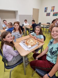 Uczniowie klasy piatej siedza przy stolikach na szkolnej stołówce. Na pierwszym planie widać cztery dziewczynki siedzące przy wspólnym stoliku, na którym leży pizza. Dziewczynki uśmiechają się do zdjęcia.