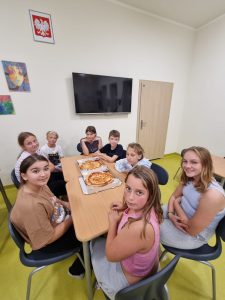 Uczniowie klasy piątej siedzą przy dwóch połączonych stołach na szkolnej stołówce. Na stołach leżą dwa pudełka z pizzami.Dzieci odwracają się w strone obiektywu i uśmiechają do zdjęcia.
