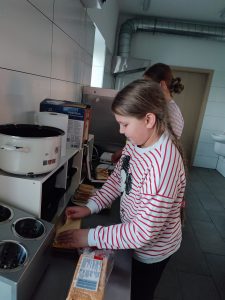 Uczennica klasy piątej podczas przygotowywania tostów w szkolnej kuchni. Dziewczynka wyciąga plasterek sera a obok leży opakowanie chleba tostowego.