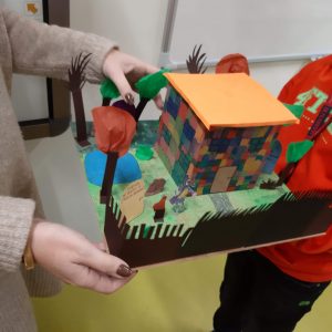 Zdjęcie makiety Akadamii Pana Kleksa wykonanej przez jednego z uczniów. Na makiecie ustawiono kolorowy budynek z płaskim dachem a w okół niego ustawiono drzewa z papieru i kreptu. Na zdjęciu widać jak uczeń trzymamakietę, a Paninauczycielka stojąca na przeciwko jej dotyka dłońmi.