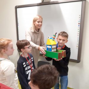 Uczeń trzyma w dłoniach wykonana przez siebie makietę Akademii Pana Kleksa. Stojąca obok nauczycielka pomaga mu ją podtrzymać. Na przeciwko stoją uczniowie klasy i przyglądają się makiecie.