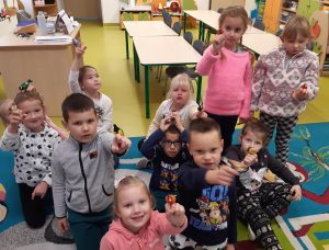 Uczniowie oddziału przedszkolnego stoją w grupie w swojej klasie. Dzieci wyciągają przed siebie kolorowe figurki zwierzątek i ptaszków.