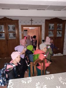 Uczniowie stoją w pomieszczeniu z drewnianymi, przeszklonymi szafami. Dzieci maja ubrane kurtki i czapki.