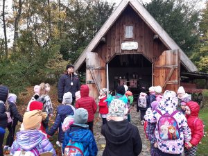 Uczniowie oddziału przedszkolnego stoją przed drewnianym domkiem ustawionym na terenie Muzeum Etnograficznego. Obok domku stoi Pan przewodnik. Dzieci odwrócone są plecami do zdjęcia.
