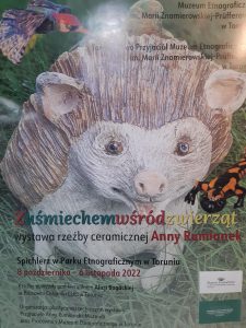 Zdjęcie plakatu przedstawiającego wykonanego z frewna jeża siedzącego wśród traw. W prawym górnym rogu napis: :Muzeum Etnograficzne im. Marii Znamierskiej-Prufferow w Toruniu. Towarzystwo Przyjaciół Muzeum Etnograficznego im. Marii Znamierskiej - Pruffer w Toruniu" Pod podobizną jeża napis: "Z uśmiechem wśród zwierząt wystawa rzeźby ceramicznej Anny Rumianek Spichlerz w Parku Etnograficznym w Toruniu 8 października - 6 listopada 2222 Rzeźby powstałe pod kierownictwem Alicji Bogackiej w Pracowni Ceramiki CUD w Toruniu" a pod spodem dopisane : "Organizacja plastyczna i techniczna wystawy. Przyjaciele Anny Rumianek i Muzeum oraz Pracownicy Muzeum Etnograficznego w Toruniu. "