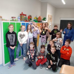 Uczniowie klasy czwartej pozują do zdjęcia na tle kolorowych szkolnych szafek na których ustawiono makiety akademii Pana Kleksa przez nich wykonane. Część uczniów podnosi w górę dłonie i układa je w różne formy.