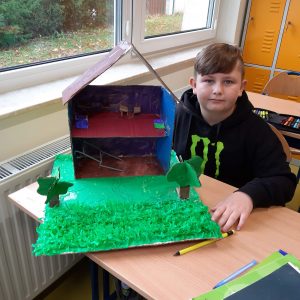 Uczeń klasy czwartej siedzi przy stoliku, na którym ustawiona jest wykonana przez niego makieta Akademii Pana Kleksa. Makieta przedstawia przekrój dwupiętrowego budynku ze spadzistym dachem.