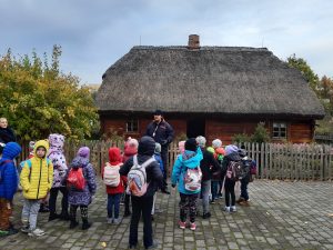 Uczniowie oddziału przedszkolnego stoją przed drewnianym domkiem ze strzechą ustawionym na terenie Muzeum Etnograficznego. Obok domku stoi Pan przewodnik. Dzieci odwrócone są plecami do zdjęcia.