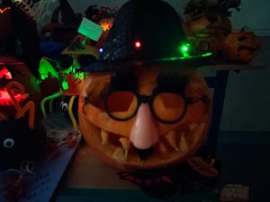 Zdjęcie dyni przygotowanej na konkurs. Dynia ozdobiona jest okularami połączonymi ze sztucznym nosem oraz ubrana w czarny kapelusz przepasany na rądzie kolorowymi lampkami.