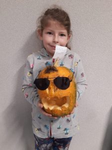 Uczennica trzyma w ręku wykonaną przez siebie dynię z szeroko otwartymi ustami i okularami przeciwsłonecznymi.